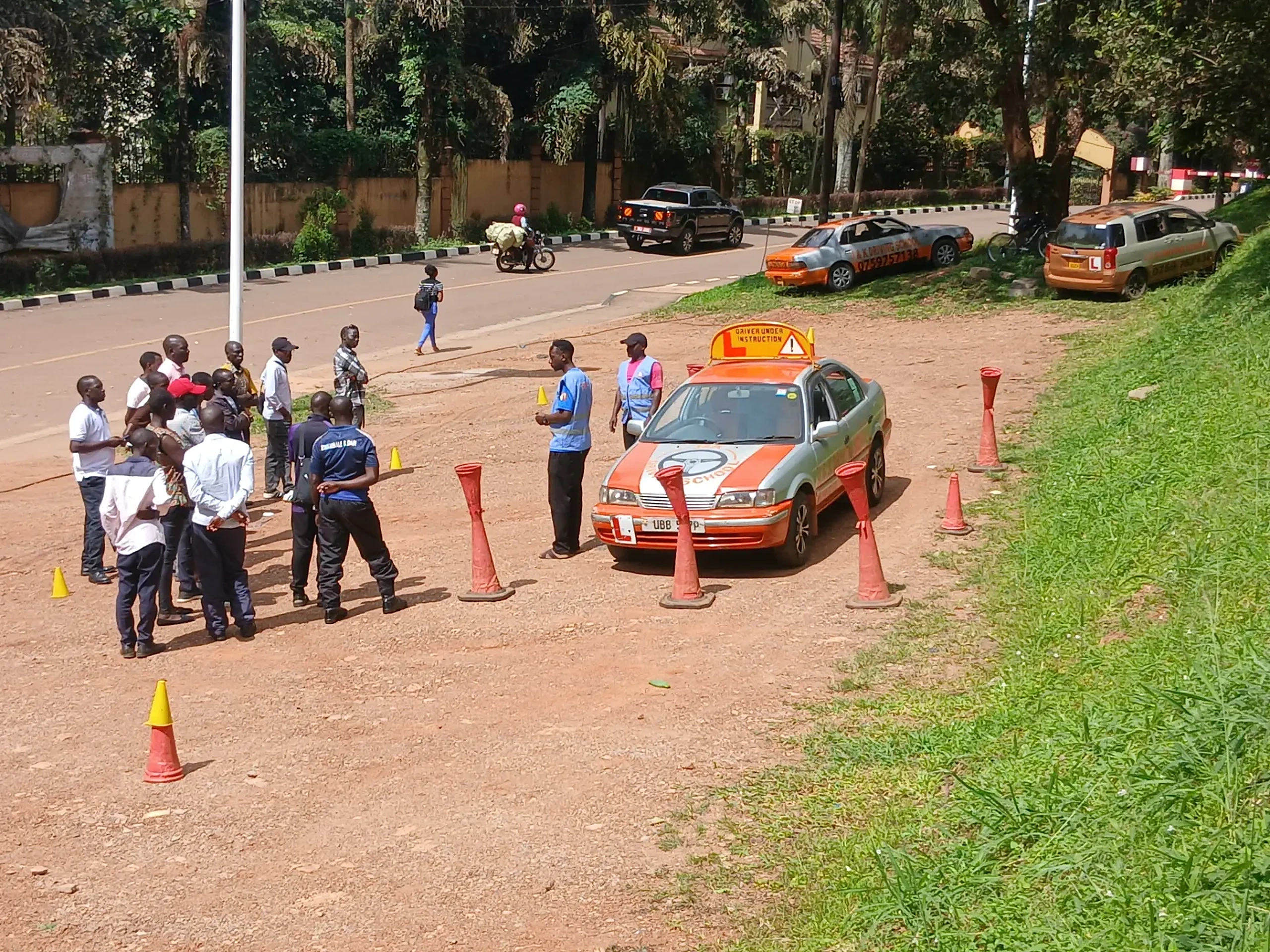 J&K Driving School learners and instructors around a Driver Under Instruction training car on a coned practice range in Kampala